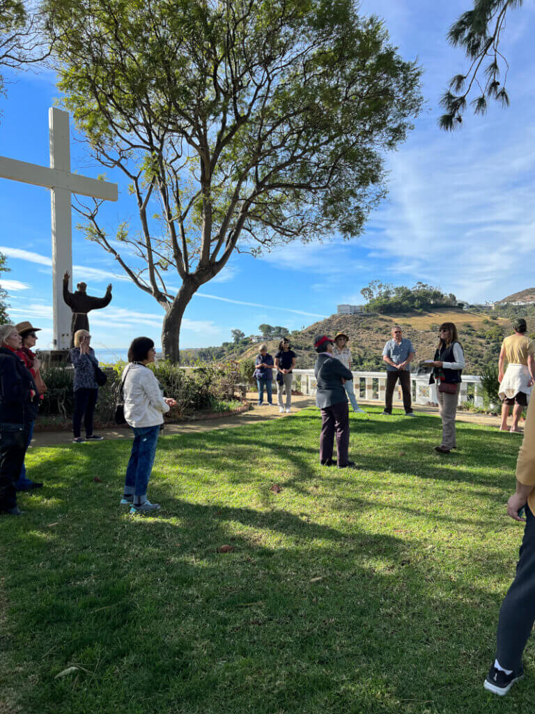 Garden Club members learning about the retreat in a grassy area under the shade of a tree with a large cross on the left.